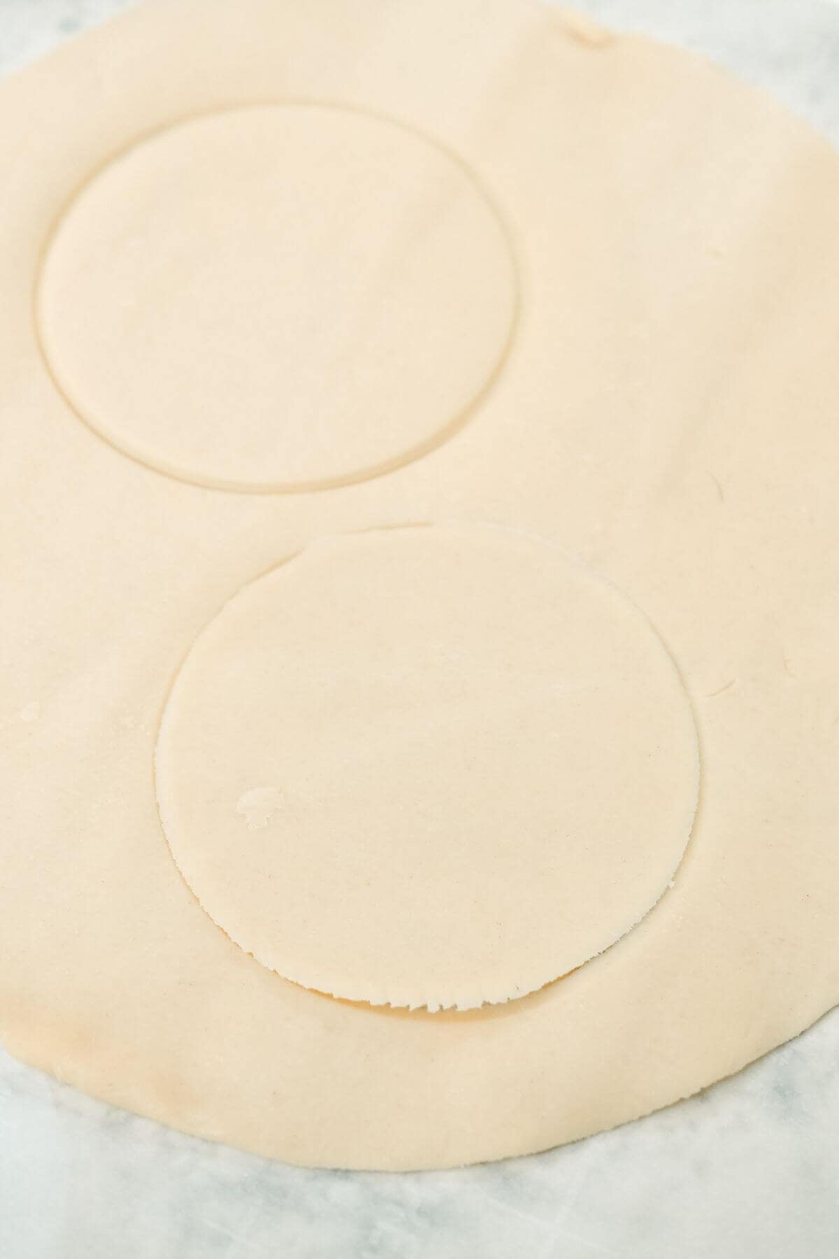 Two large circular shapes are cut out from a rolled piece of pie dough on a flat surface.