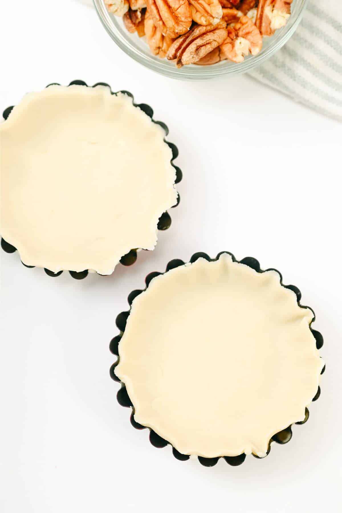 Two small tart pans lined with unbaked pie crust dough. A bowl of chopped nuts is in the background.