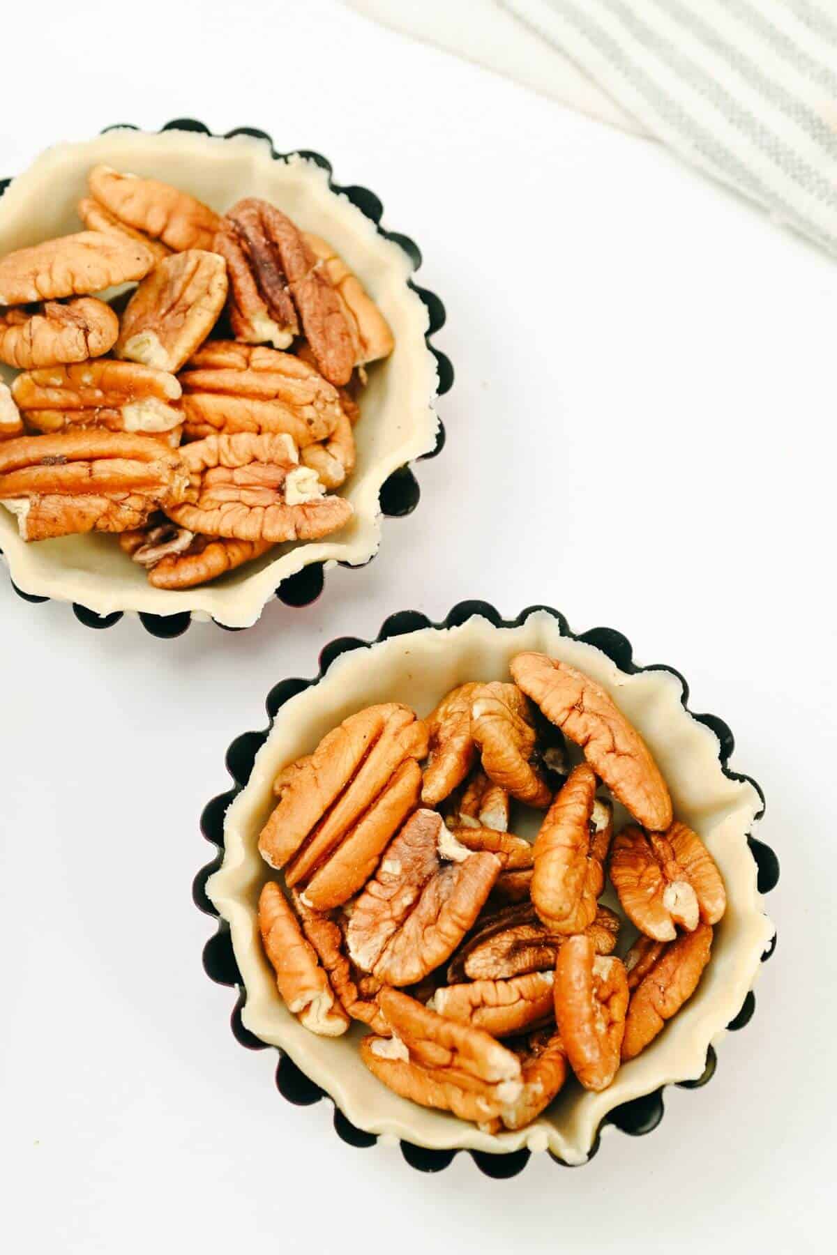 Two scalloped-edge tart pans are filled with pecan nuts, placed on a white surface.