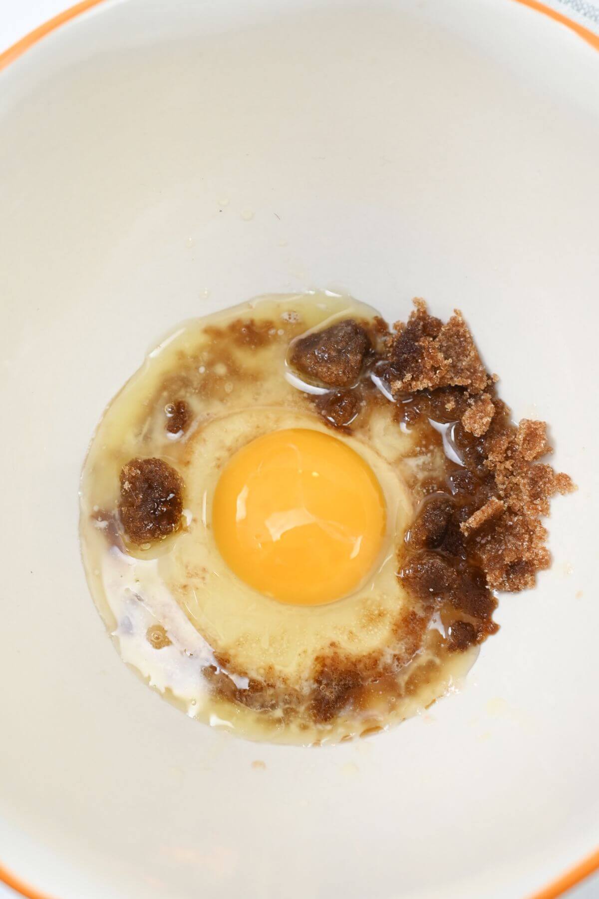 A mixing bowl containing an egg, brown sugar, and other ingredients preparing to be mixed.