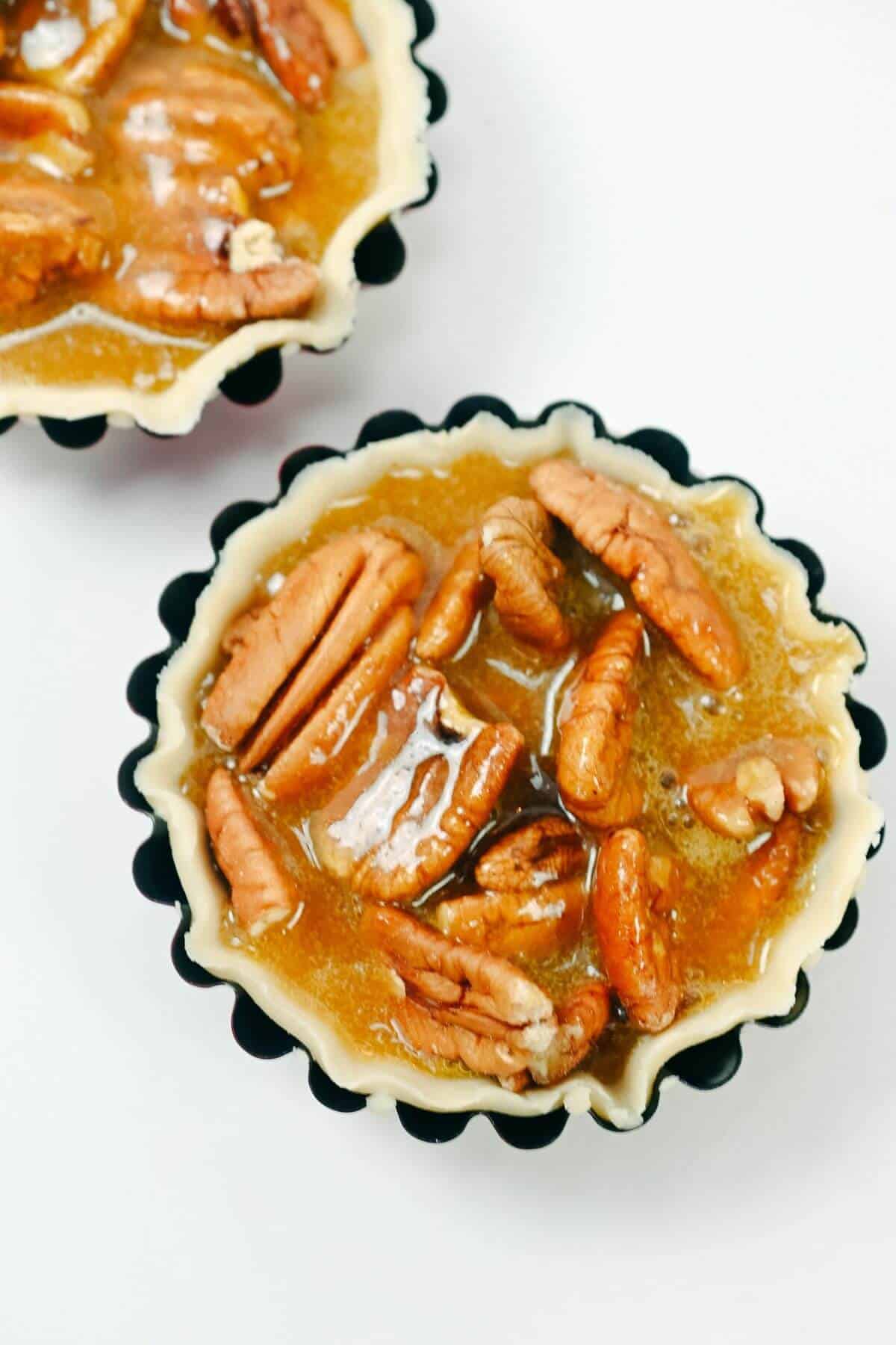 Two unbaked mini pecan pies in black fluted tart pans filled with a brown sugar and pecan mixture, placed on a white surface.