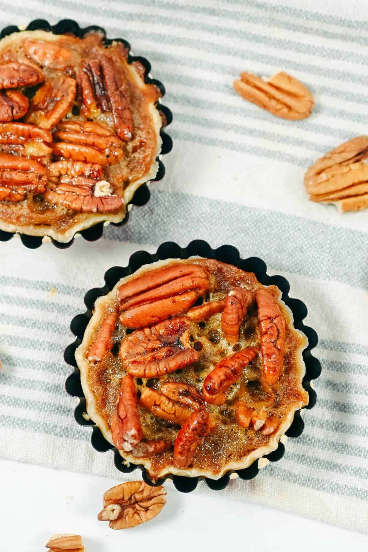 Two pecan tarts in black tart pans are placed on a striped cloth, surrounded by scattered pecan halves.