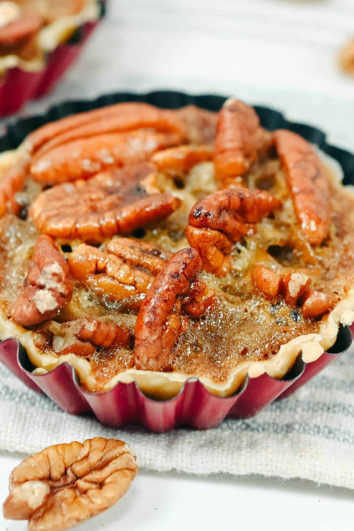 A close-up view of a mini pecan pie with a golden-brown filling, topped with whole pecan halves. The tart is in a red fluted baking dish, and a few pecan pieces are scattered nearby.