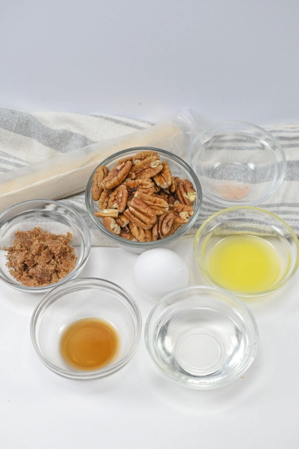 Various baking ingredients including pecans, brown sugar, vanilla extract, butter, egg, vinegar, and a pinch of salt, displayed in small glass bowls, with a rolled pie dough in the background.
