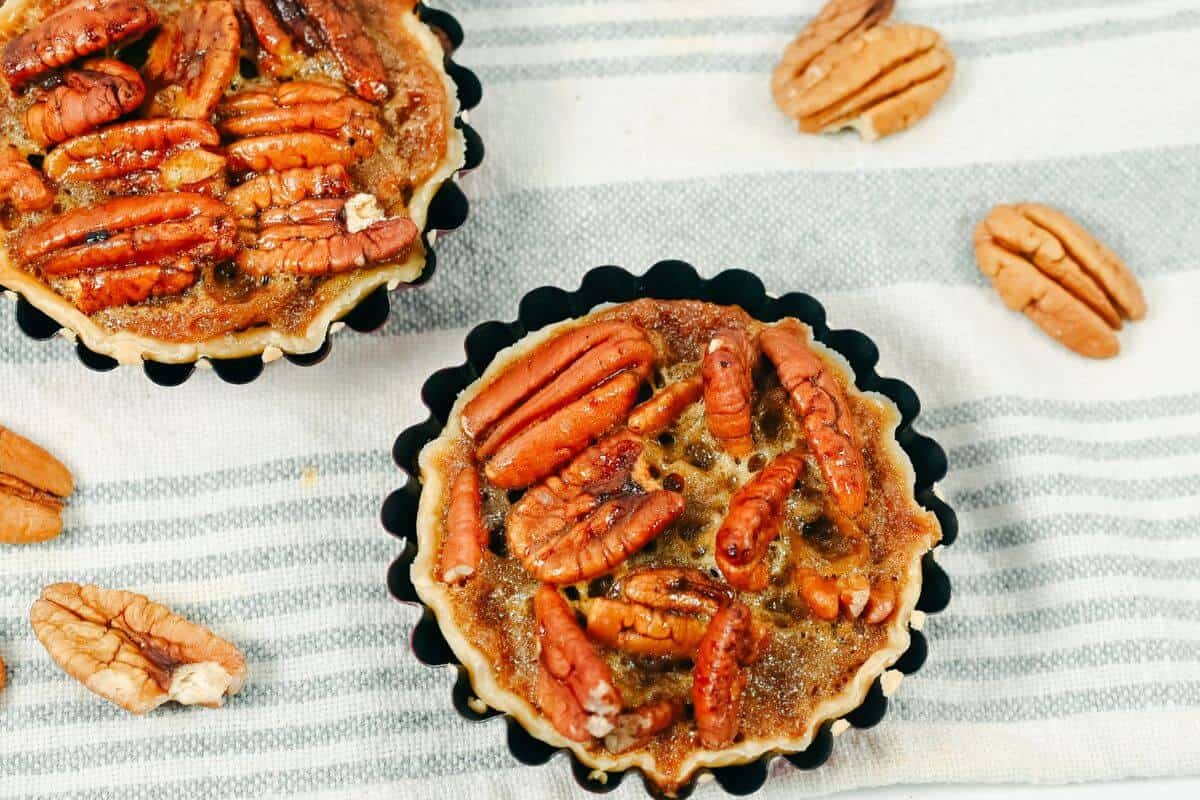 Two mini pecan pies in fluted tart pans sit on a striped cloth, surrounded by several whole pecans.