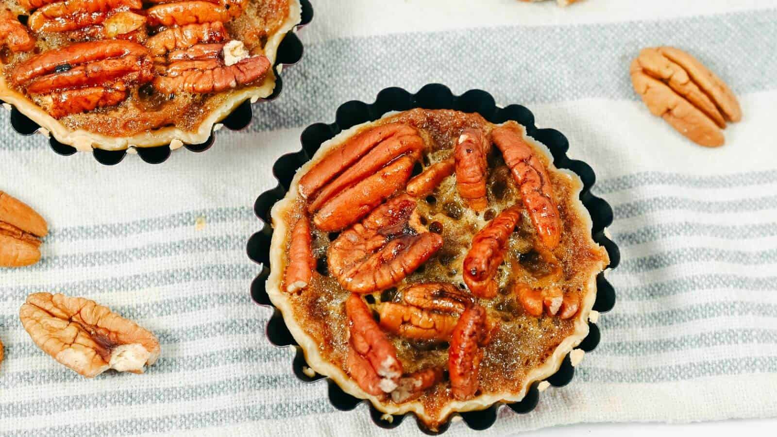 Two mini pecan pies in black tart pans rest on a white and gray striped cloth, surrounded by scattered pecan halves.