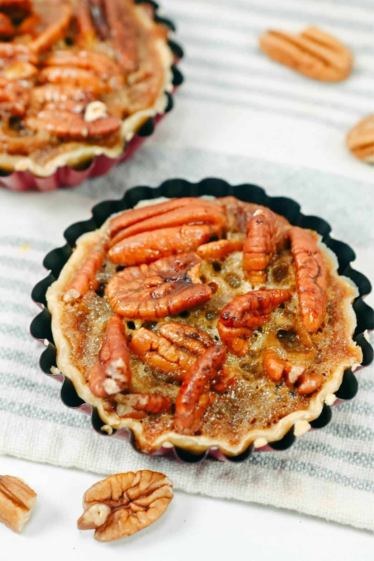 A close-up of a pecan pie in a mini fluted tart pan, topped with whole and halved pecans sitting on a striped cloth with a few loose pecans scattered around.