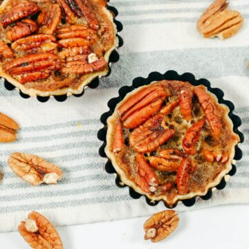 Two miniature pecan pies with golden crusts and caramelized pecans, placed on a striped cloth napkin, surrounded by scattered pecan halves.