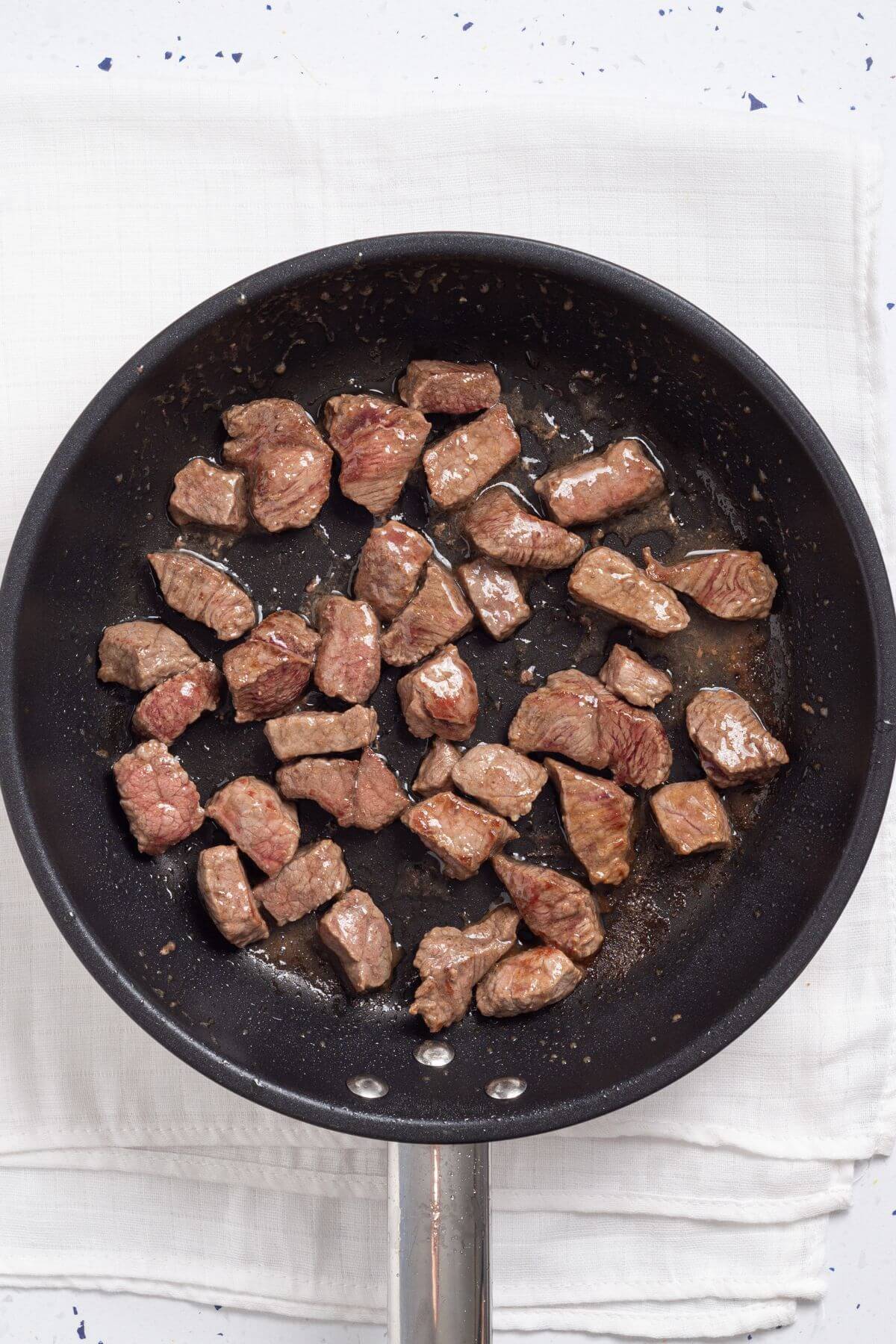 Chunks of beef being seared in a black frying pan over a white cloth.