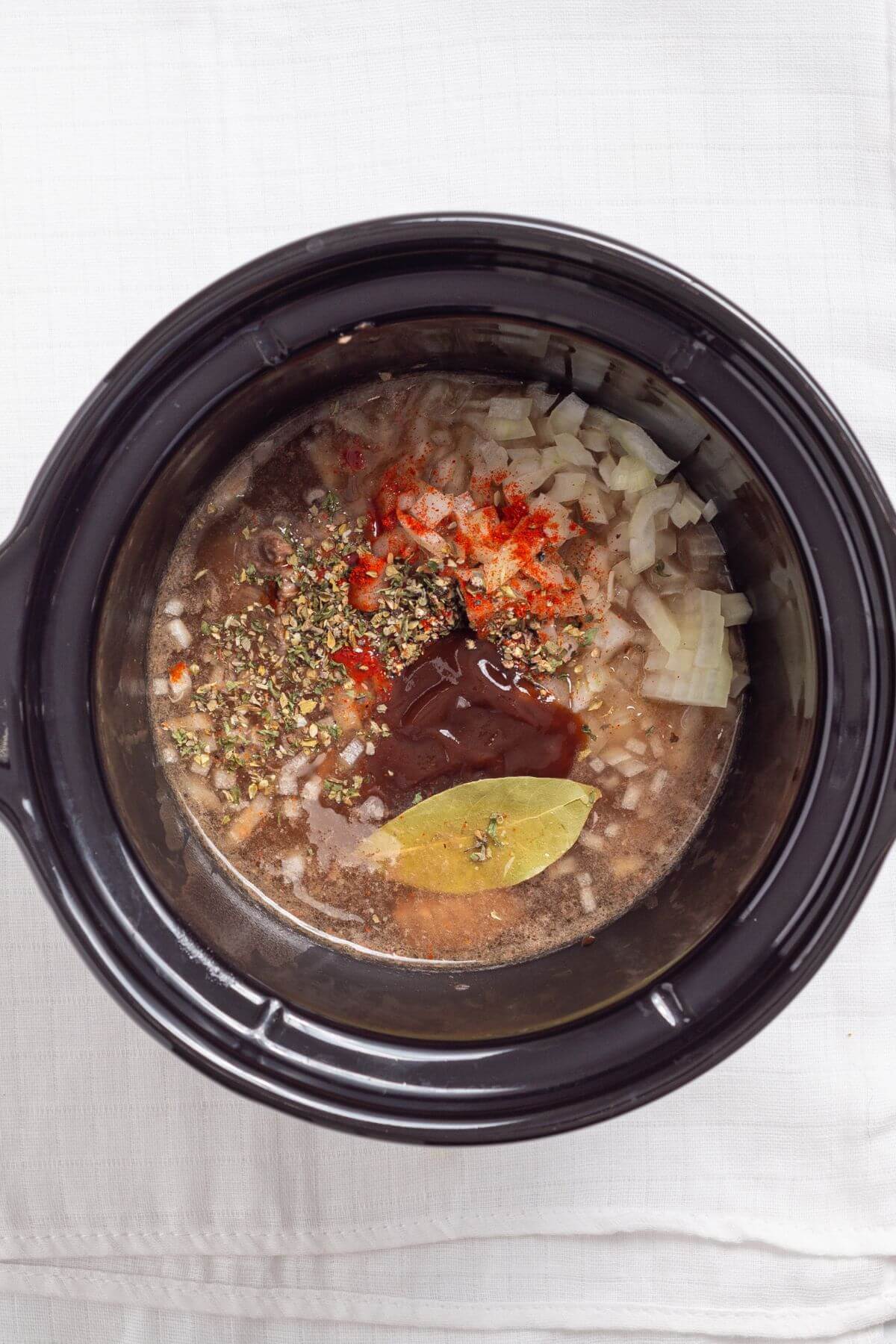 A slow cooker filled with chopped onions, spices, and sauce mix. A bay leaf rests on top. The ingredients surround a brown liquid base.