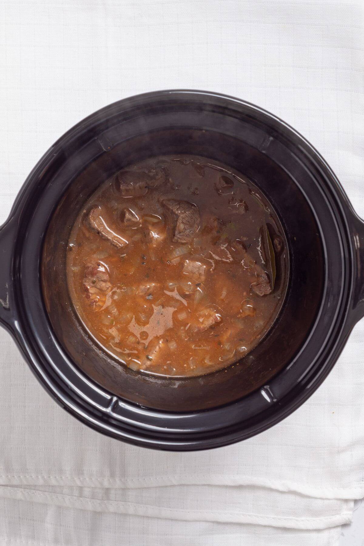 A slow cooker filled with beef stew, featuring broth, beef chunks, and vegetables, placed on a white tablecloth.