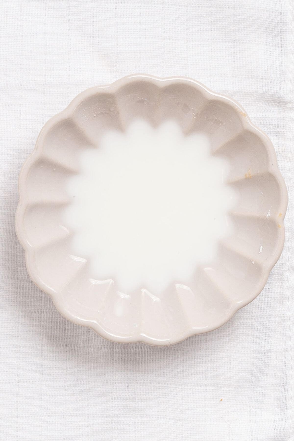 A small, empty, scalloped-edged ceramic dish with a pale color is placed on a white textured cloth.