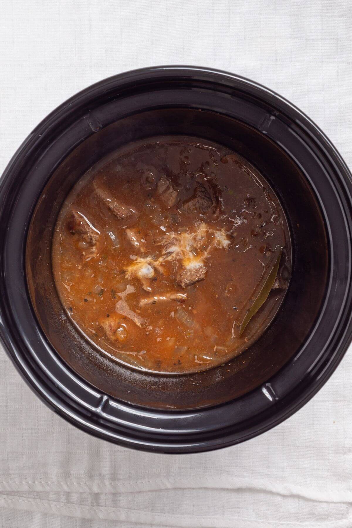 A black slow cooker containing a brown stew with chunks of meat, a bay leaf, and visible vegetables.