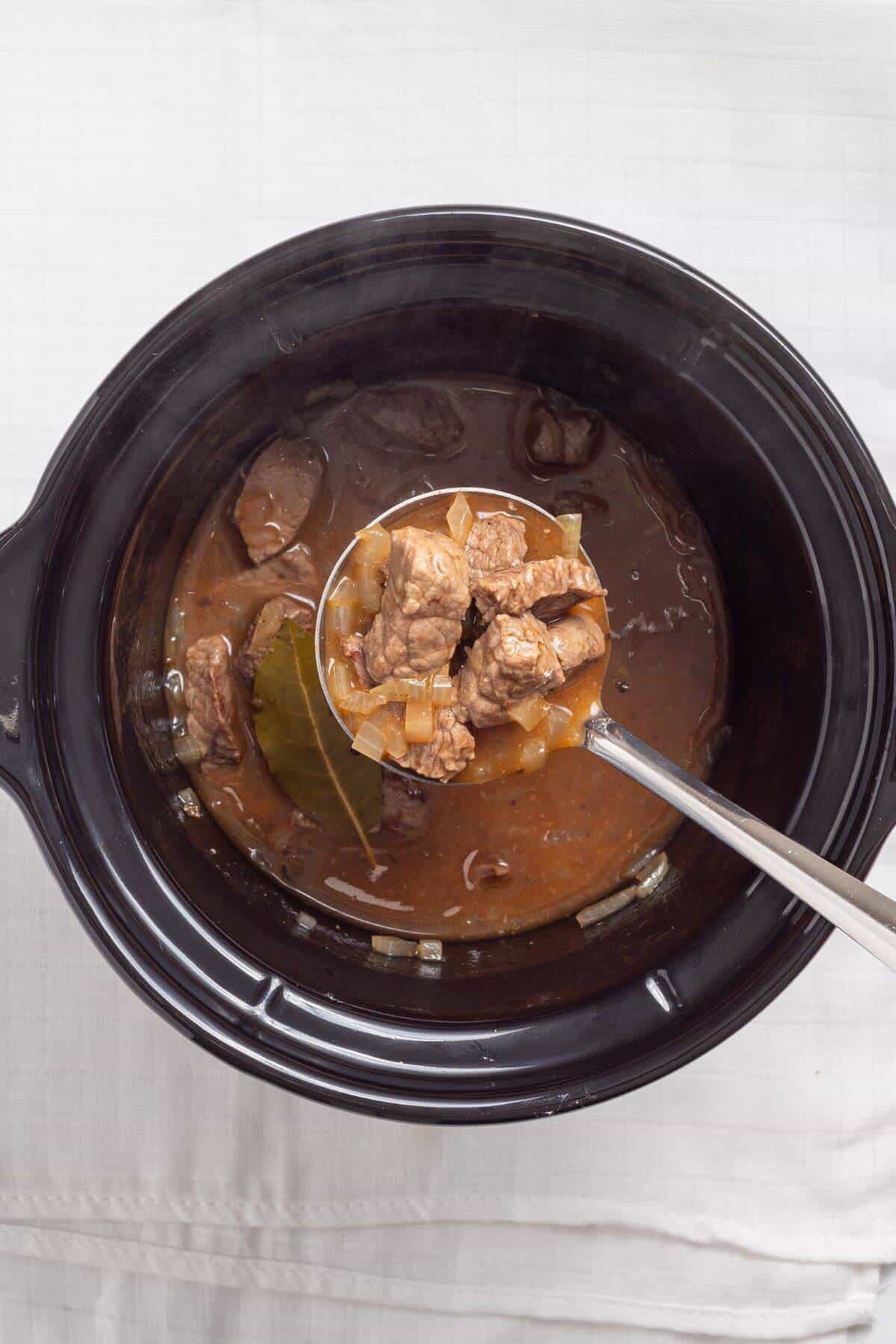 A ladleful of beef stew with chunks of meat and onions is lifted out of a black slow cooker containing the same stew.