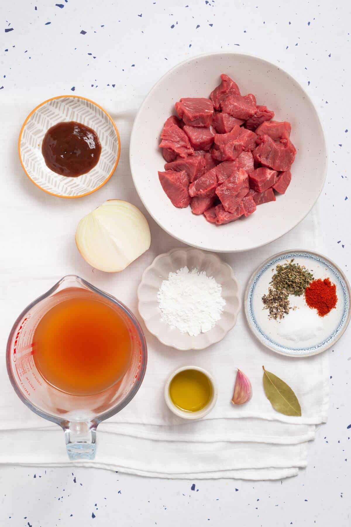 Ingredients for a recipe laid out on a light surface, including chopped beef, onion, garlic, broth, oil, sauces, spices, herbs, and flour.
