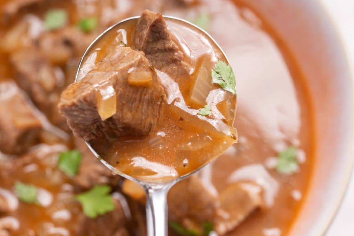 A close-up of a spoonful of beef stew with chunks of beef, onions, and garnished with green herbs in a brown broth.