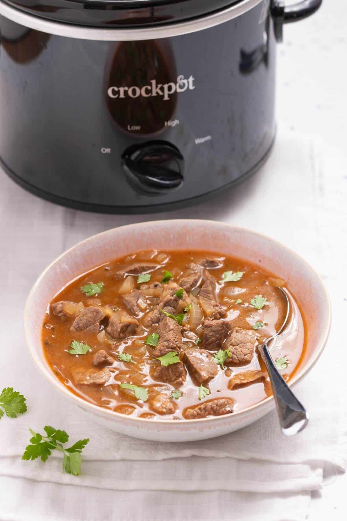 A bowl of beef stew with vegetables, garnished with cilantro, placed in front of a crockpot.