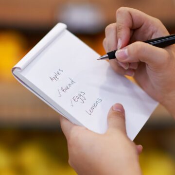A person holding a notepad and writing a grocery list that includes apples, bread, eggs, and lemons, with blurred produce in the background.