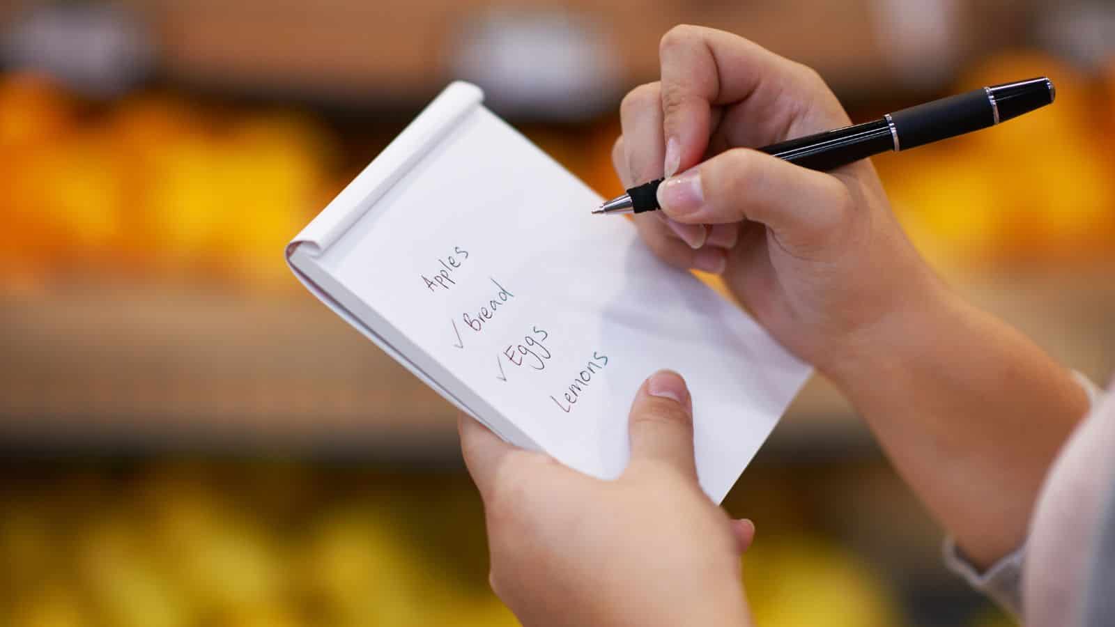 A person holding a notepad and writing a grocery list that includes apples, bread, eggs, and lemons, with blurred produce in the background.