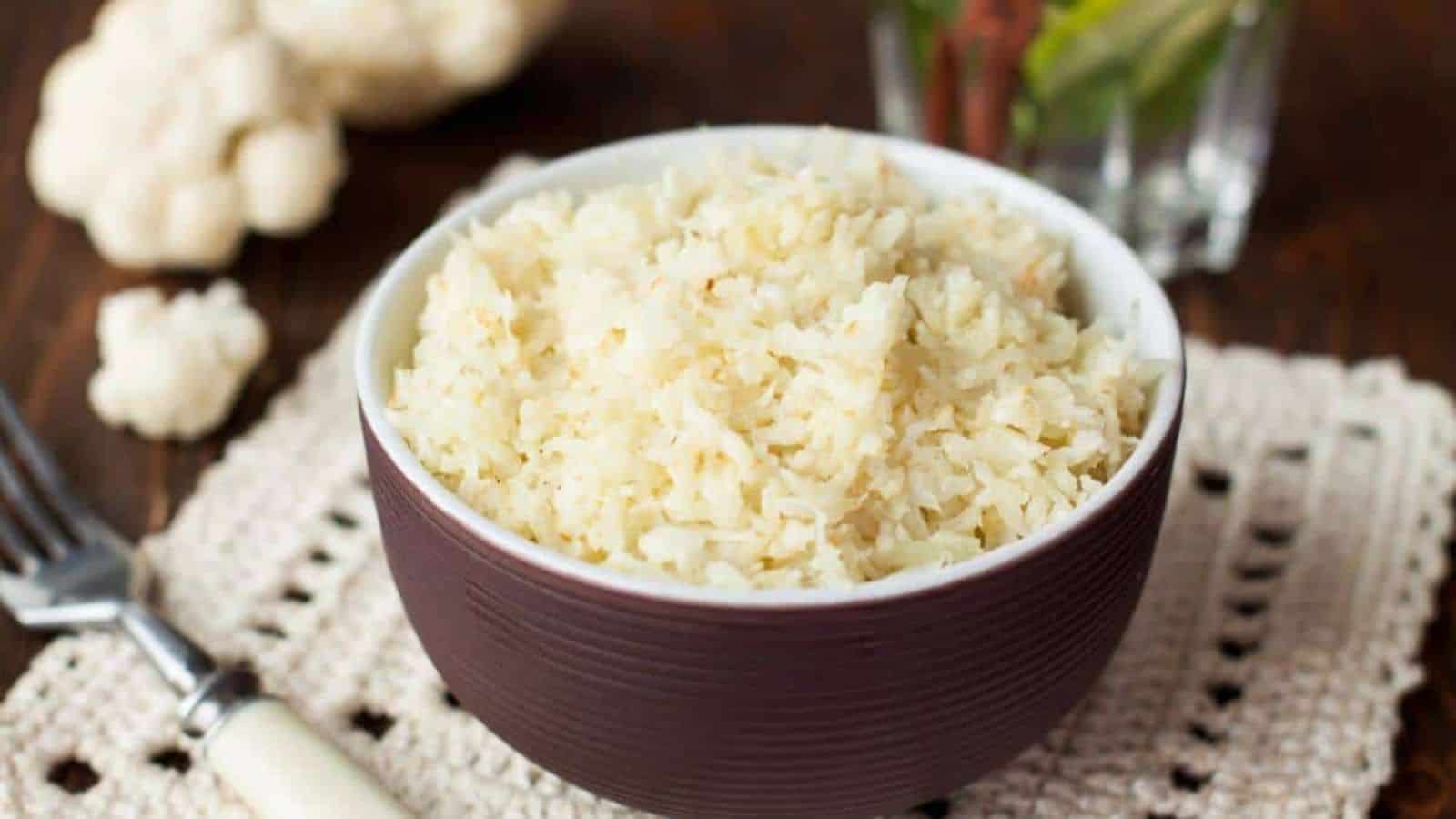 A bowl of cooked cauliflower rice is placed on a crocheted placemat beside a fork. Blurry background shows cauliflower and green leaves.