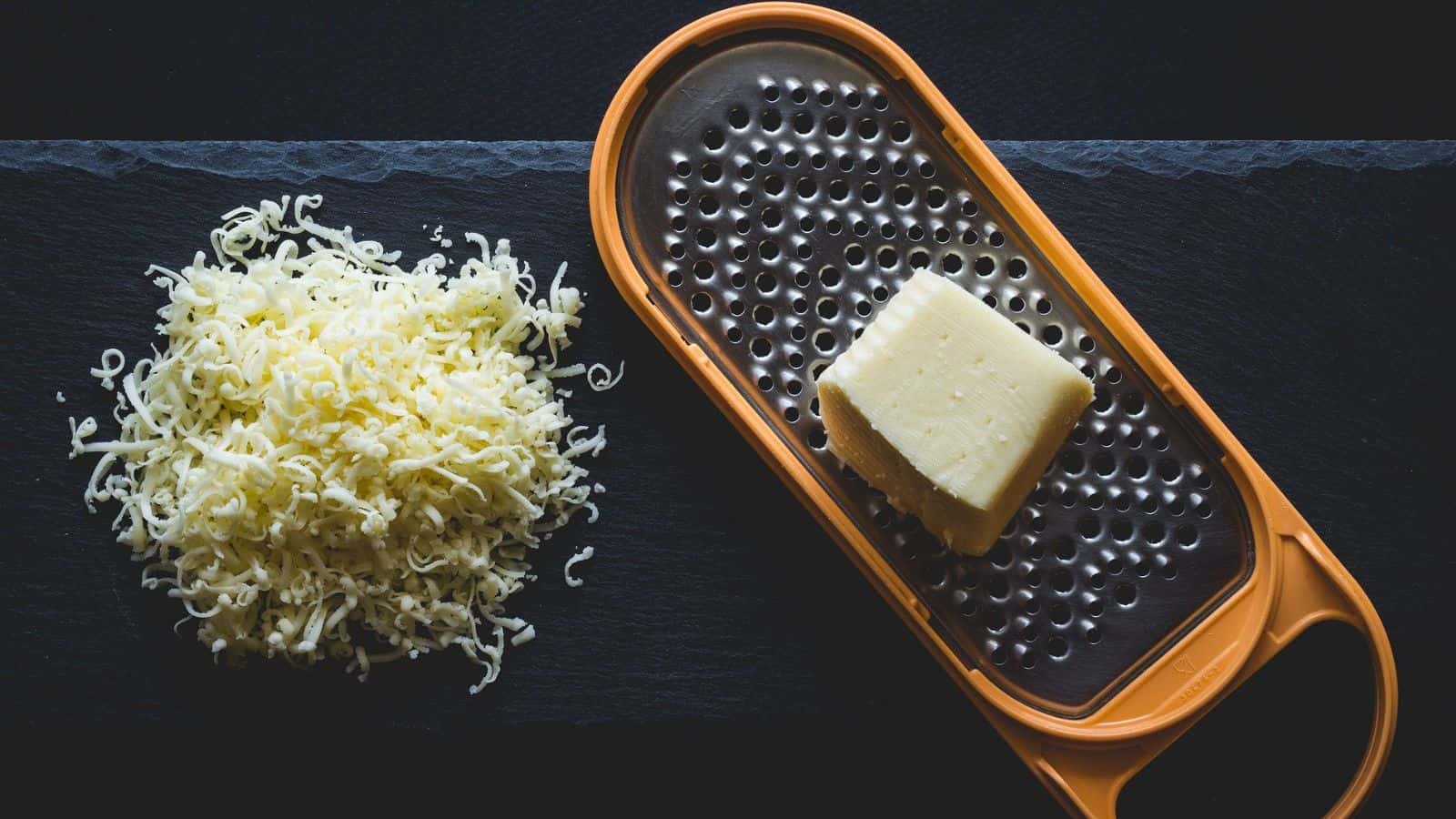 Shredded cheese is on a black surface next to an orange cheese grater with a block of cheese on it.