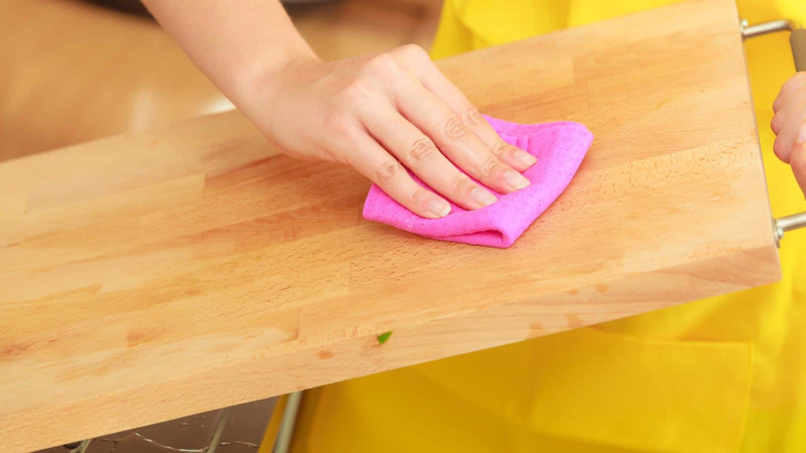 A person wearing a yellow apron is cleaning a wooden surface with a pink cloth.