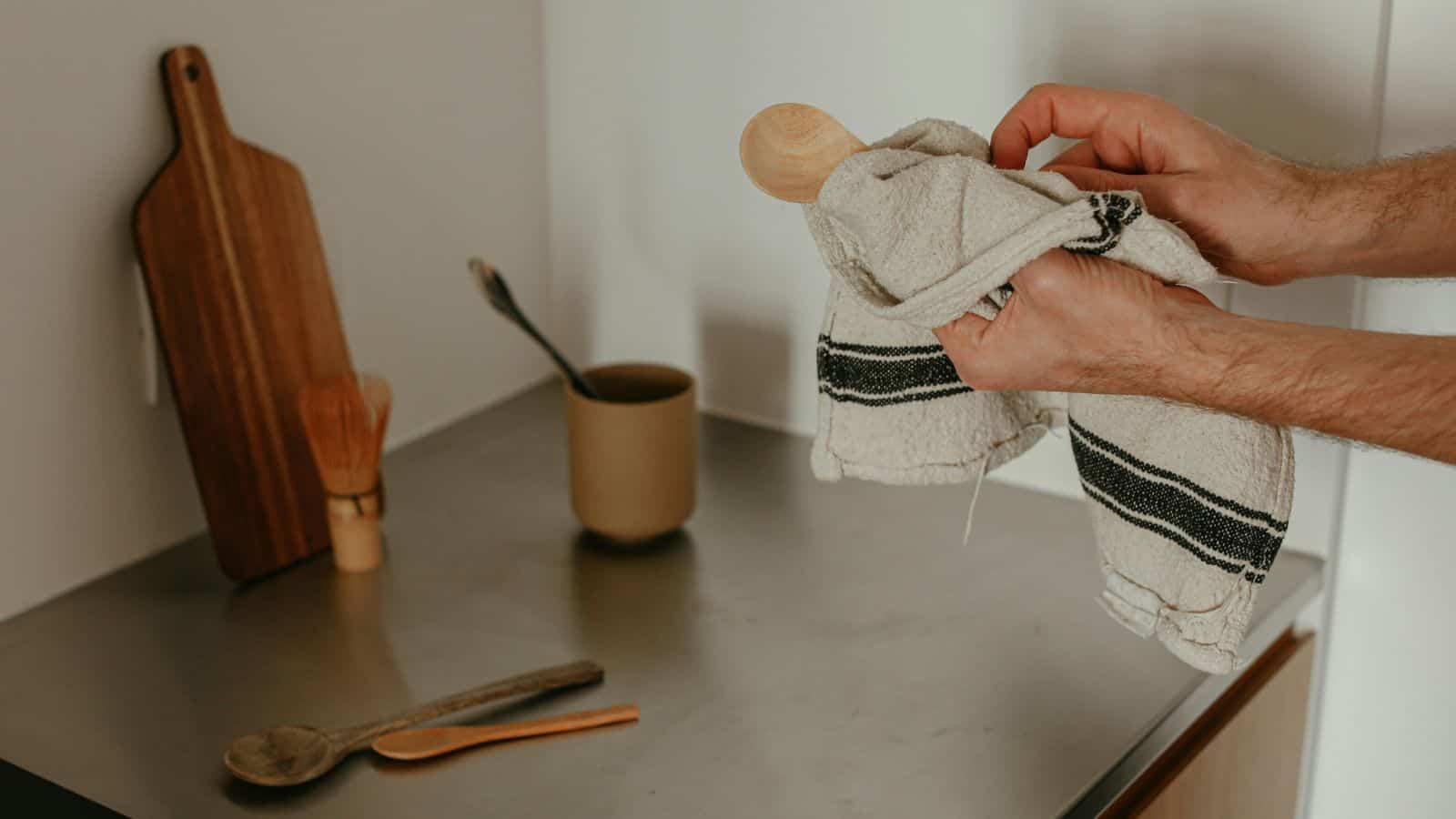 A person dries a wooden spoon with a cloth on a kitchen counter, with utensils and a cutting board in the background.