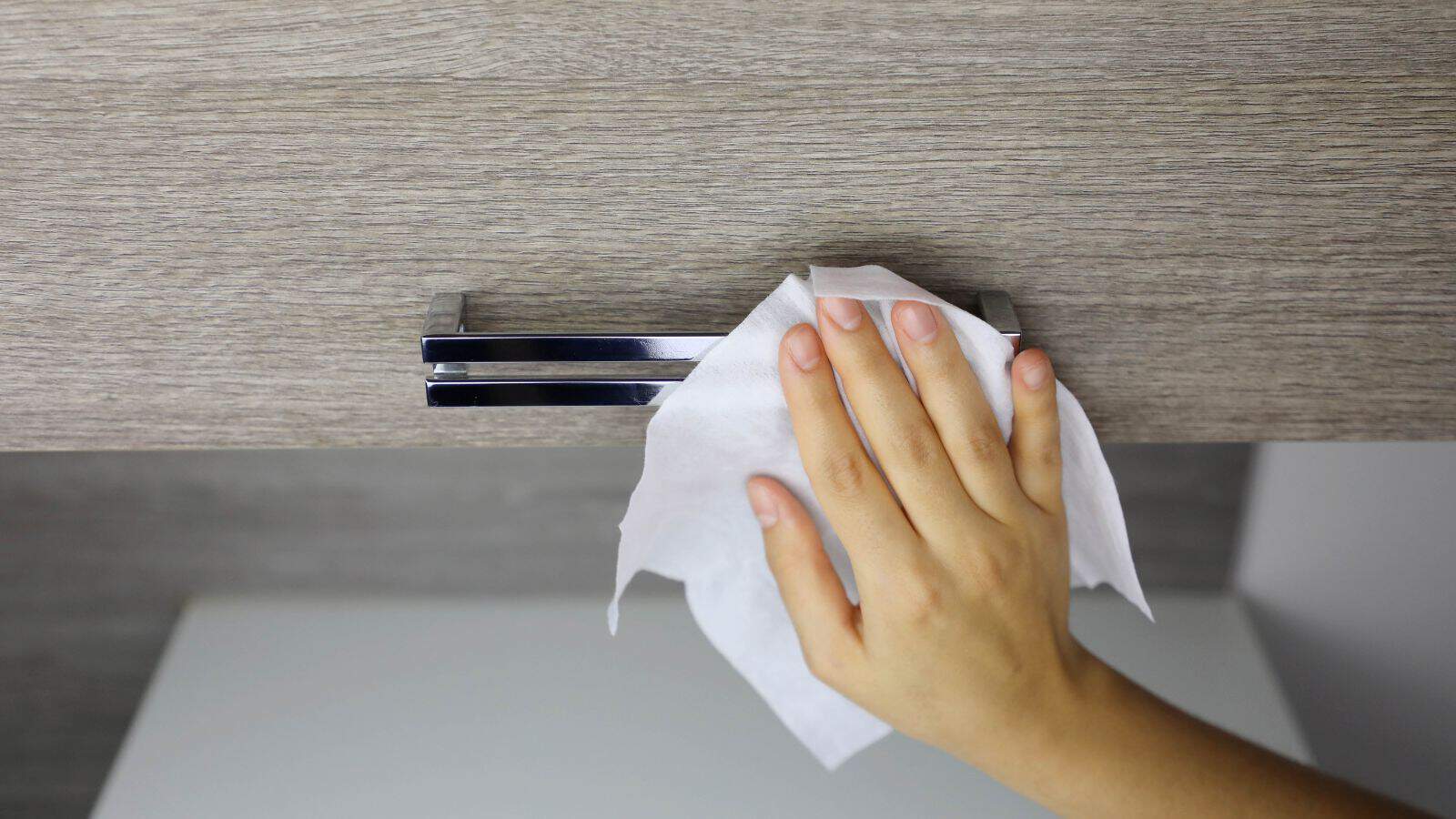A hand cleaning a wooden cabinet handle with a white cloth.