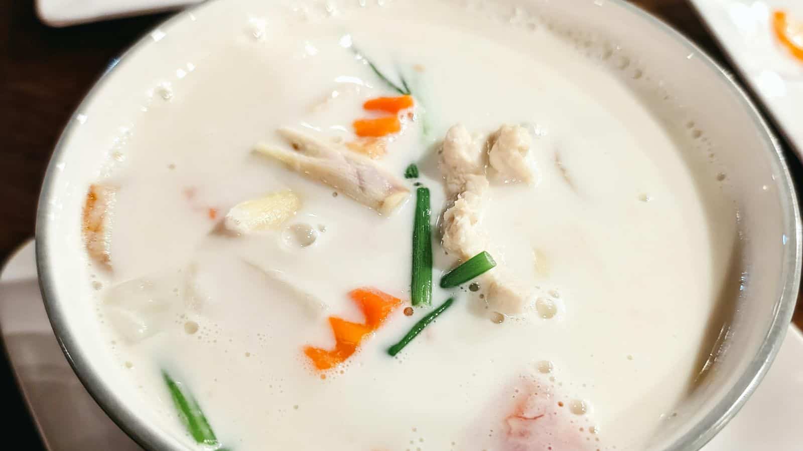 A bowl of Tom Kha Gai soup featuring a creamy coconut milk base with pieces of chicken, sliced vegetables, and herbs.