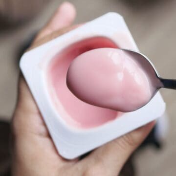 A person holds a plastic container of pink yogurt in one hand and a spoonful of the yogurt in the other hand, showcasing one of those snacks that are secretly junk food.