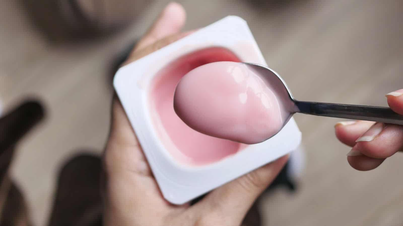 A person holds a plastic container of pink yogurt in one hand and a spoonful of the yogurt in the other hand, showcasing one of those snacks that are secretly junk food.