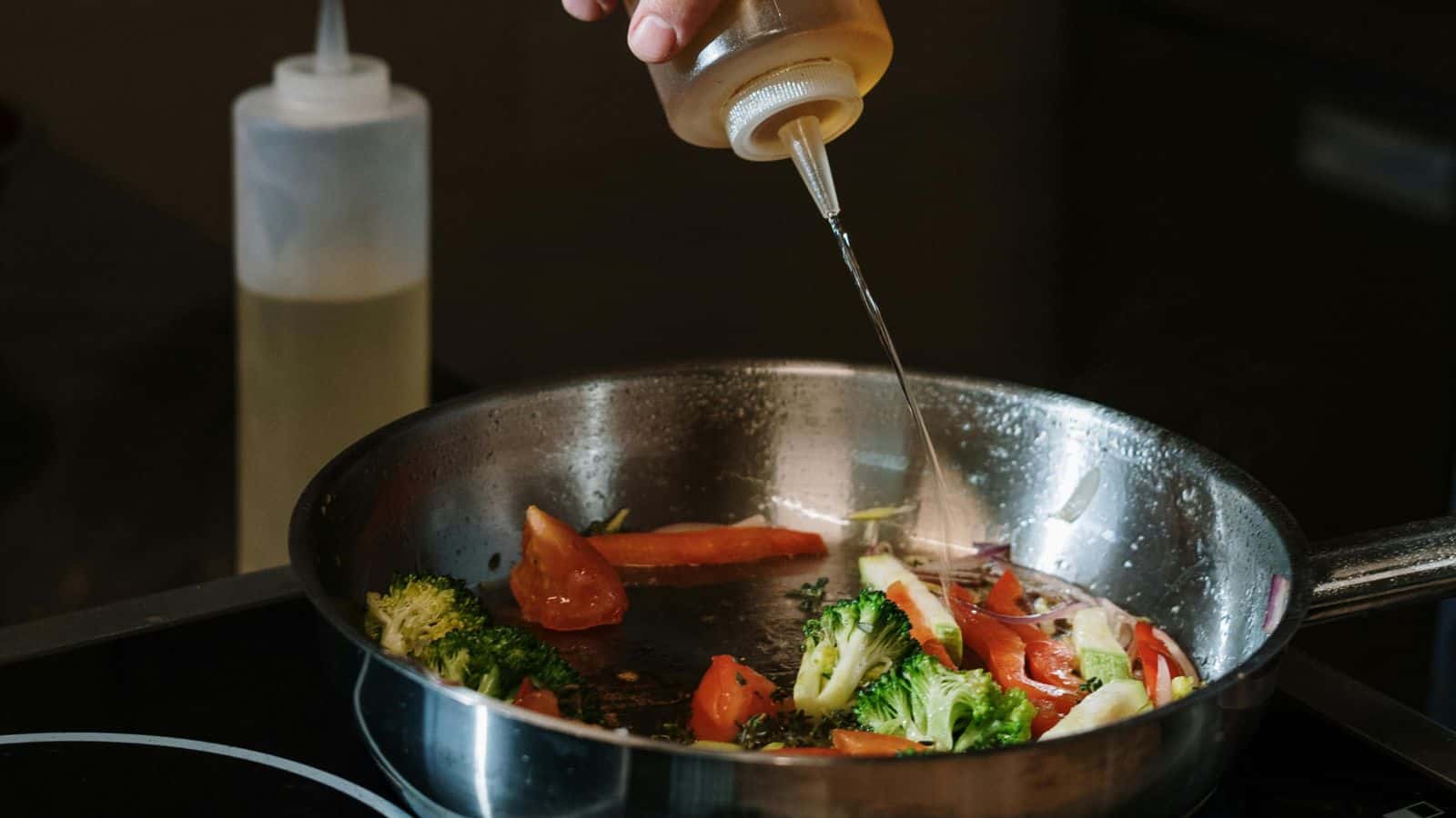 A hand pours oil from a squeeze bottle into a stainless steel pan filled with cooking vegetables, including broccoli and tomatoes, on a stove.
