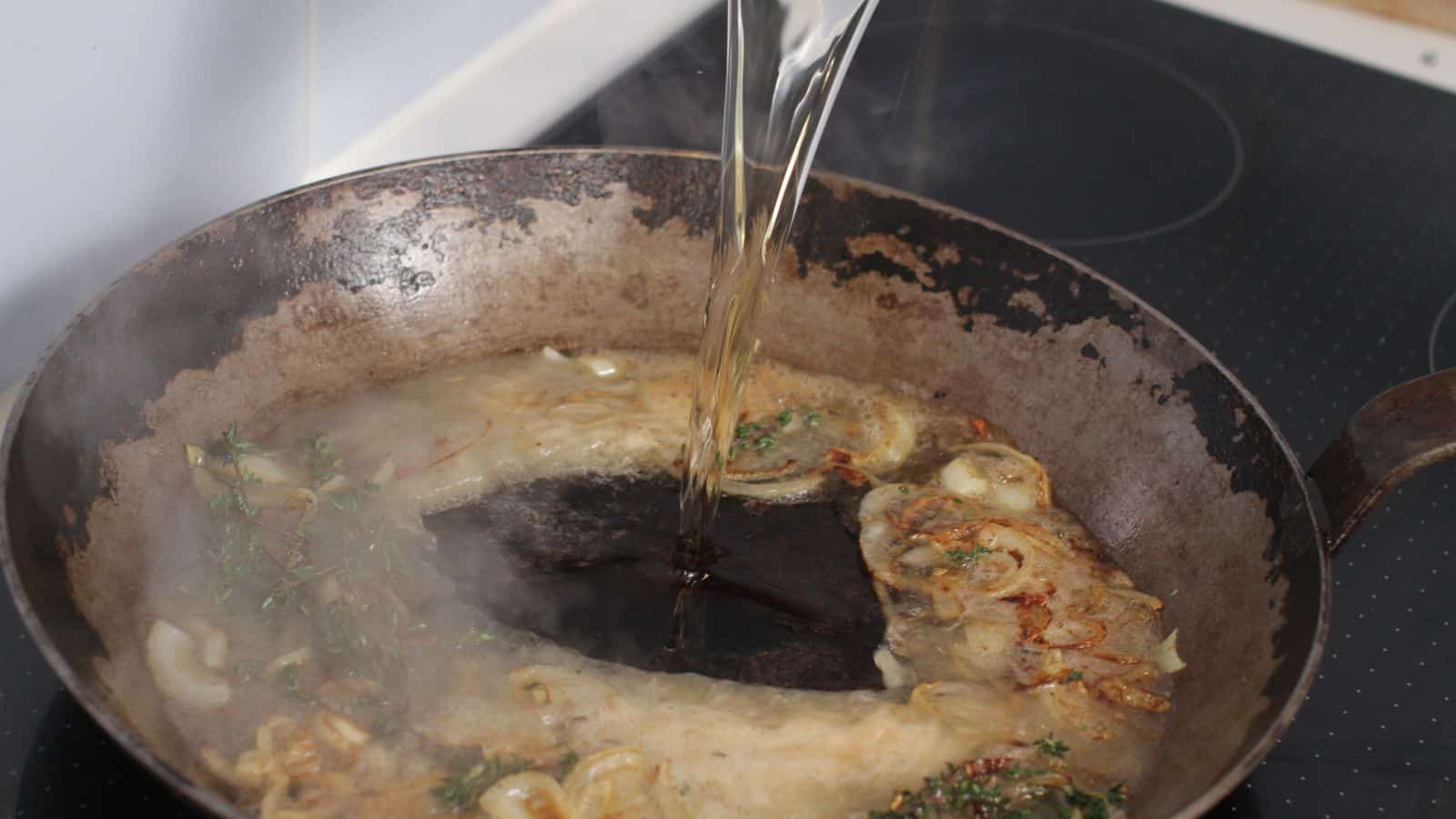 Liquid being poured into a hot skillet with sautéed onions, garlic, herbs, and browned residue, creating steam.