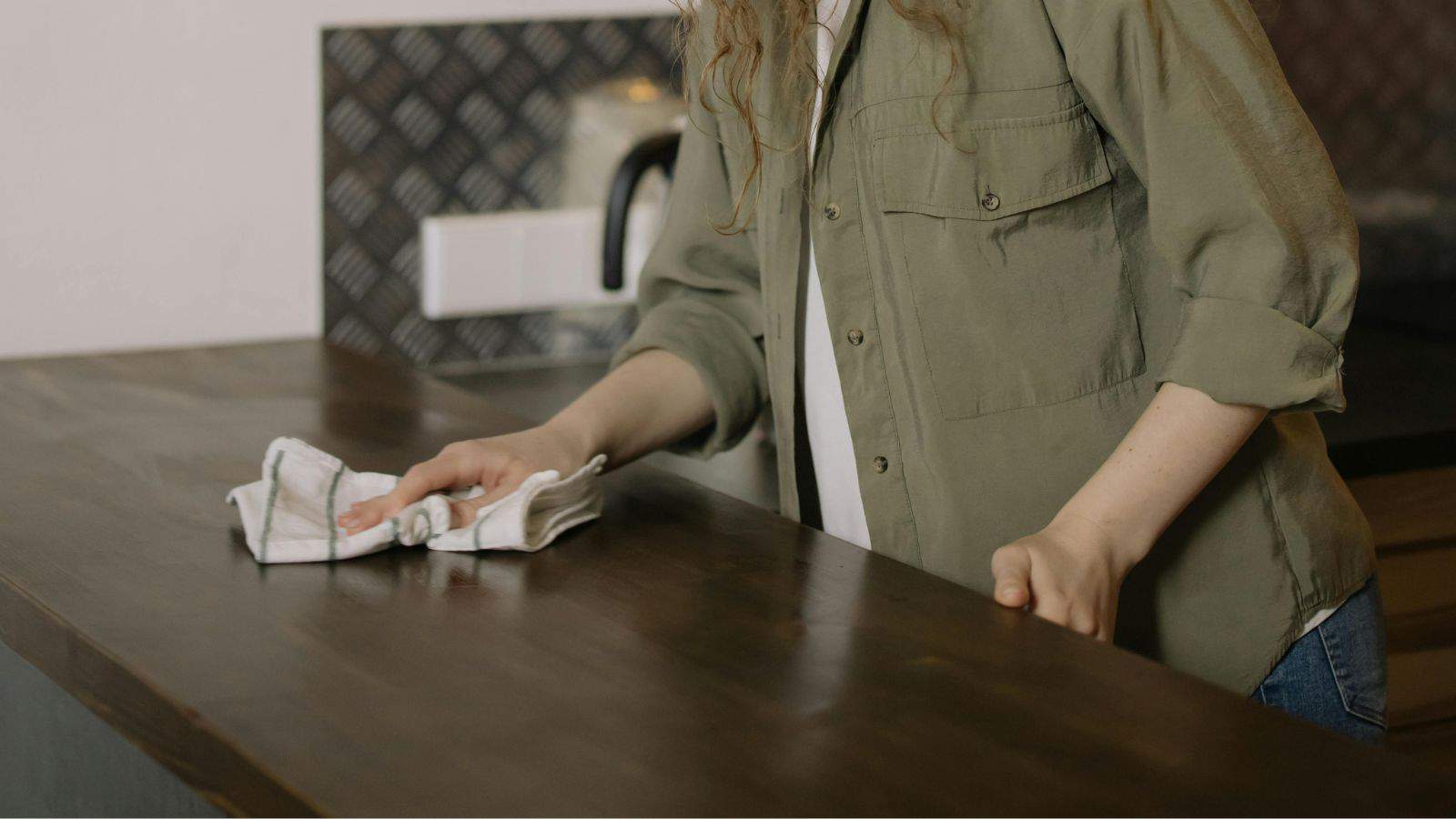 A person wearing a green shirt cleaning a countertop with a cloth in a kitchen.