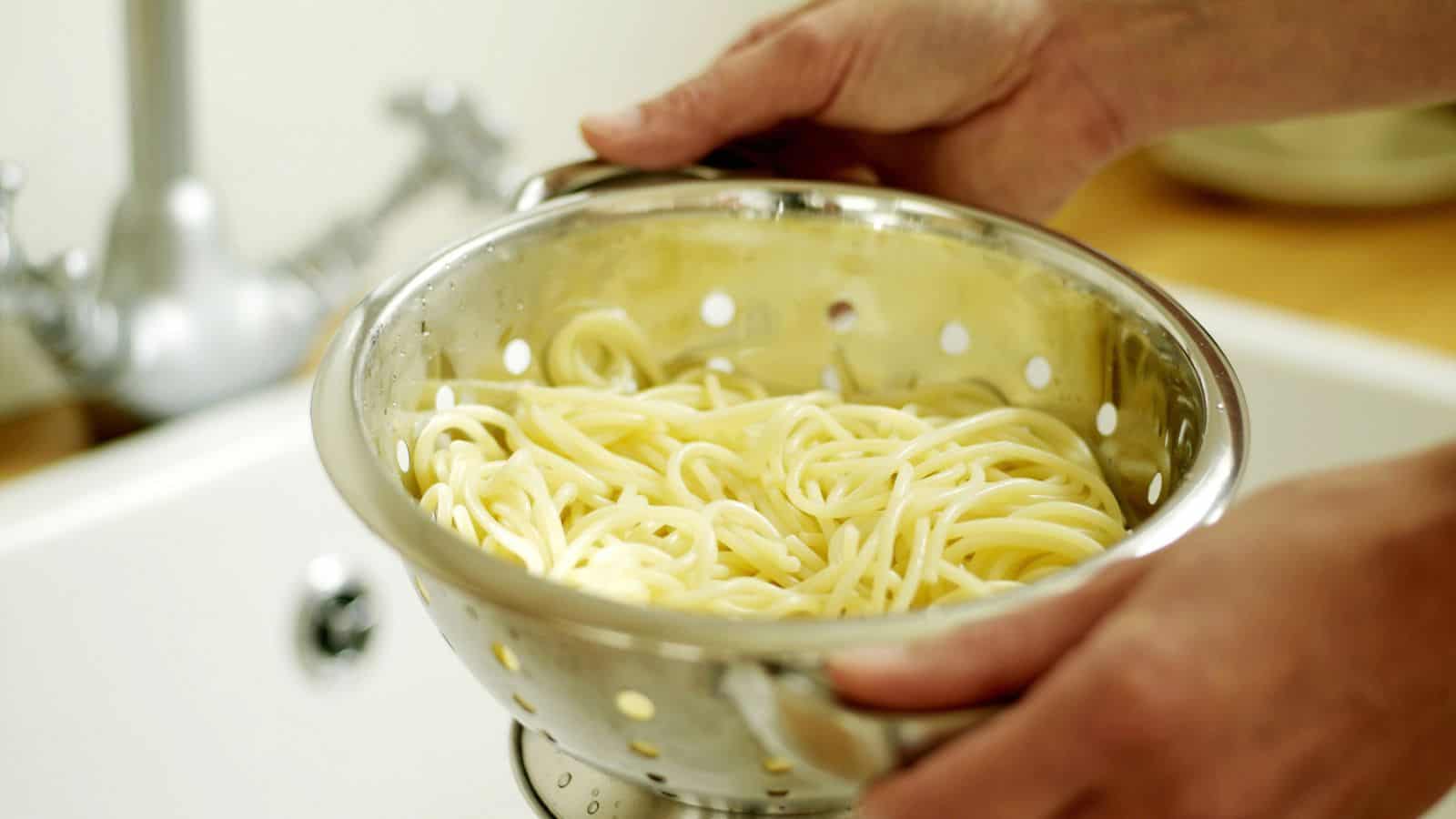 Hands holding a metal colander with cooked spaghetti over a sink.