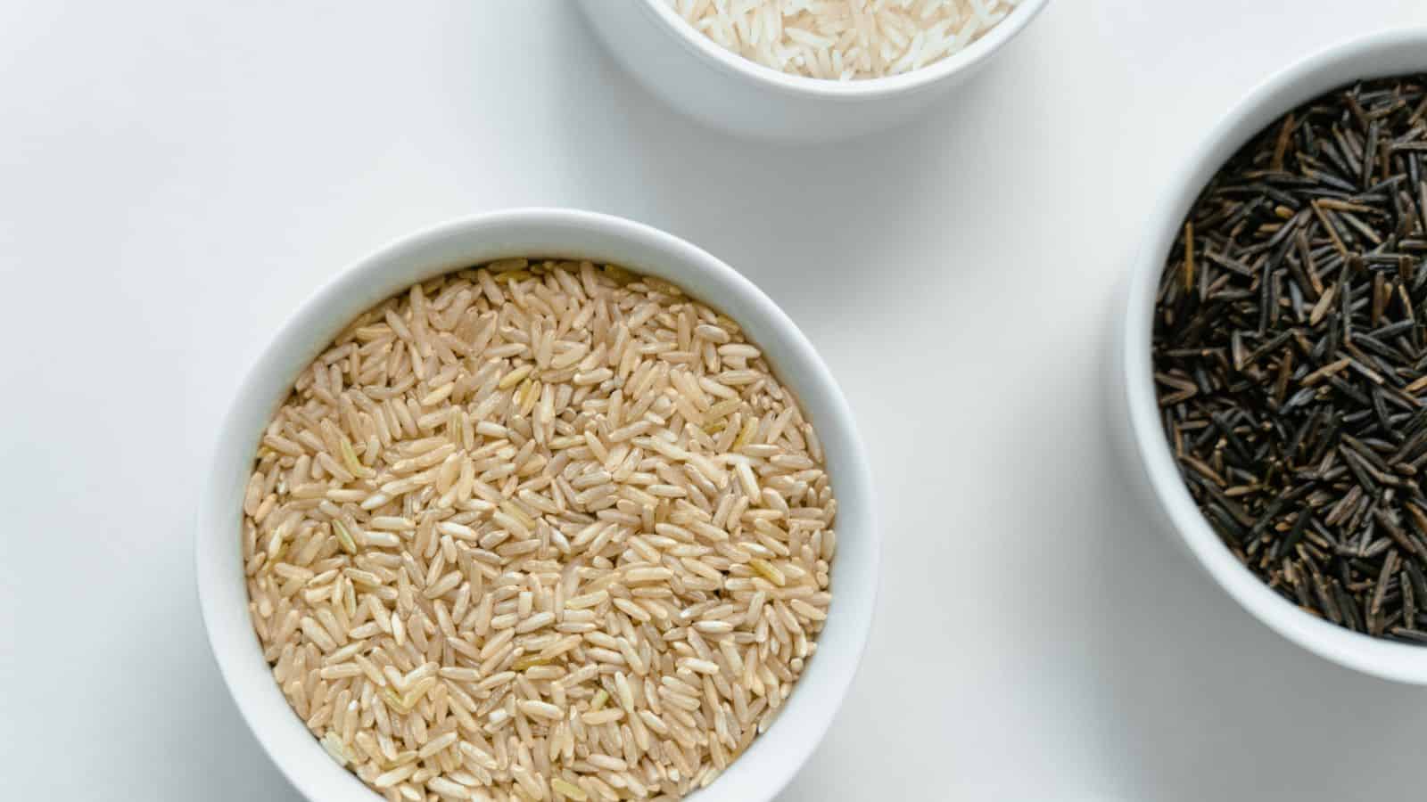 Three bowls containing different varieties of rice: brown rice in the bottom left, white rice in the top center, and wild rice in the right. The bowls are arranged on a white surface.
