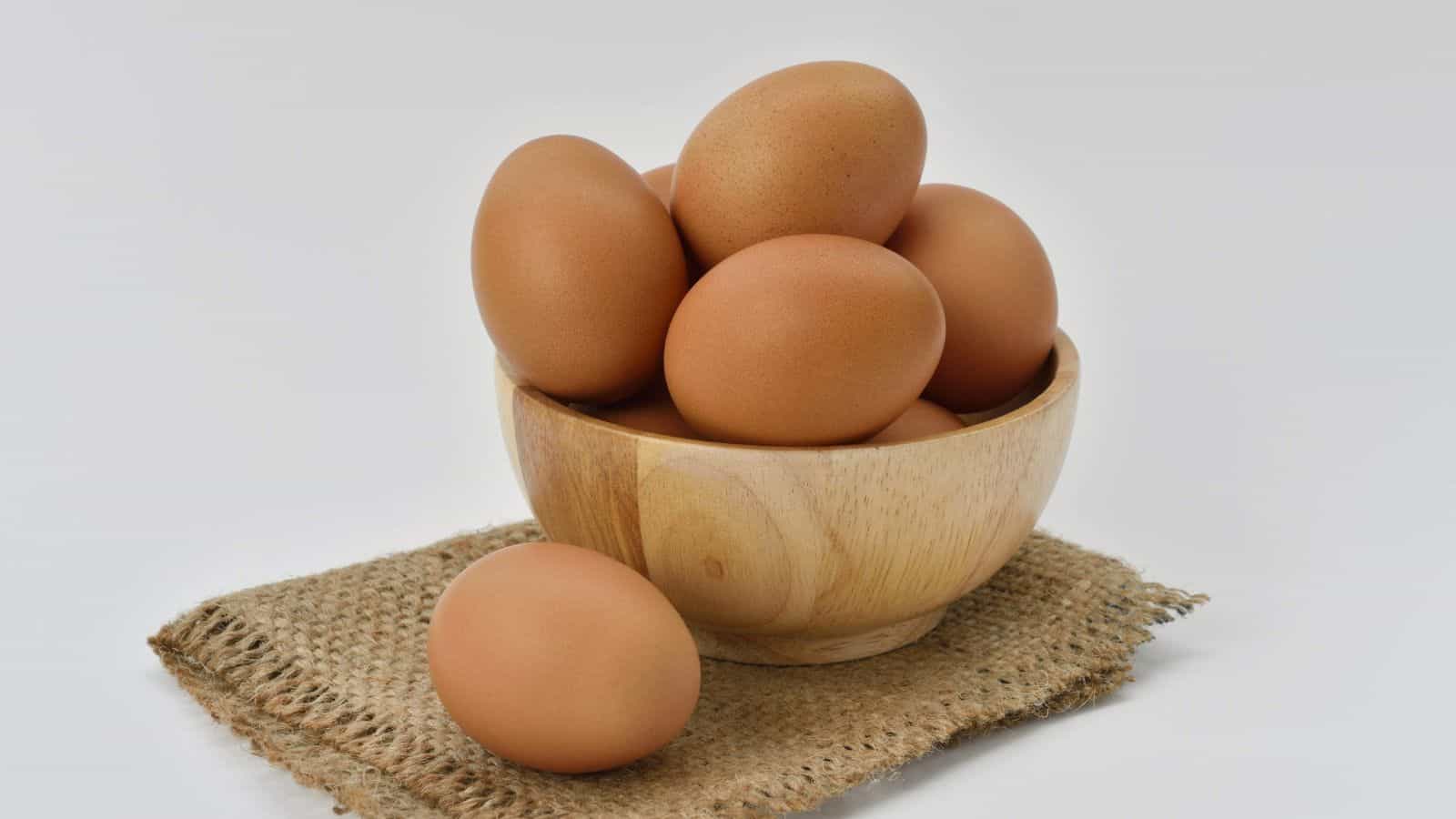 A wooden bowl filled with seven brown eggs sits on a woven mat. Two additional brown eggs are placed beside the bowl on the mat. The background is plain white.