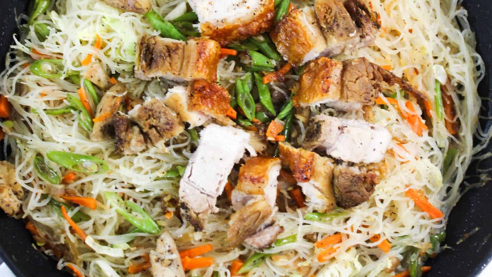 A close-up of a dish with sliced pork, vermicelli noodles, carrots, and green beans in a pan.