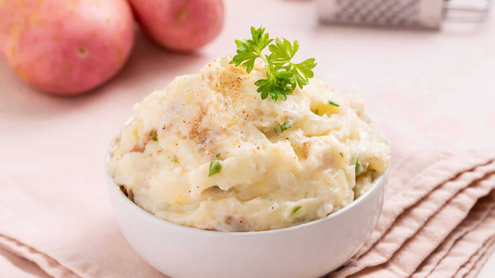 A bowl of mashed potatoes garnished with parsley, set on a pink cloth napkin, with whole potatoes and a grater in the background.