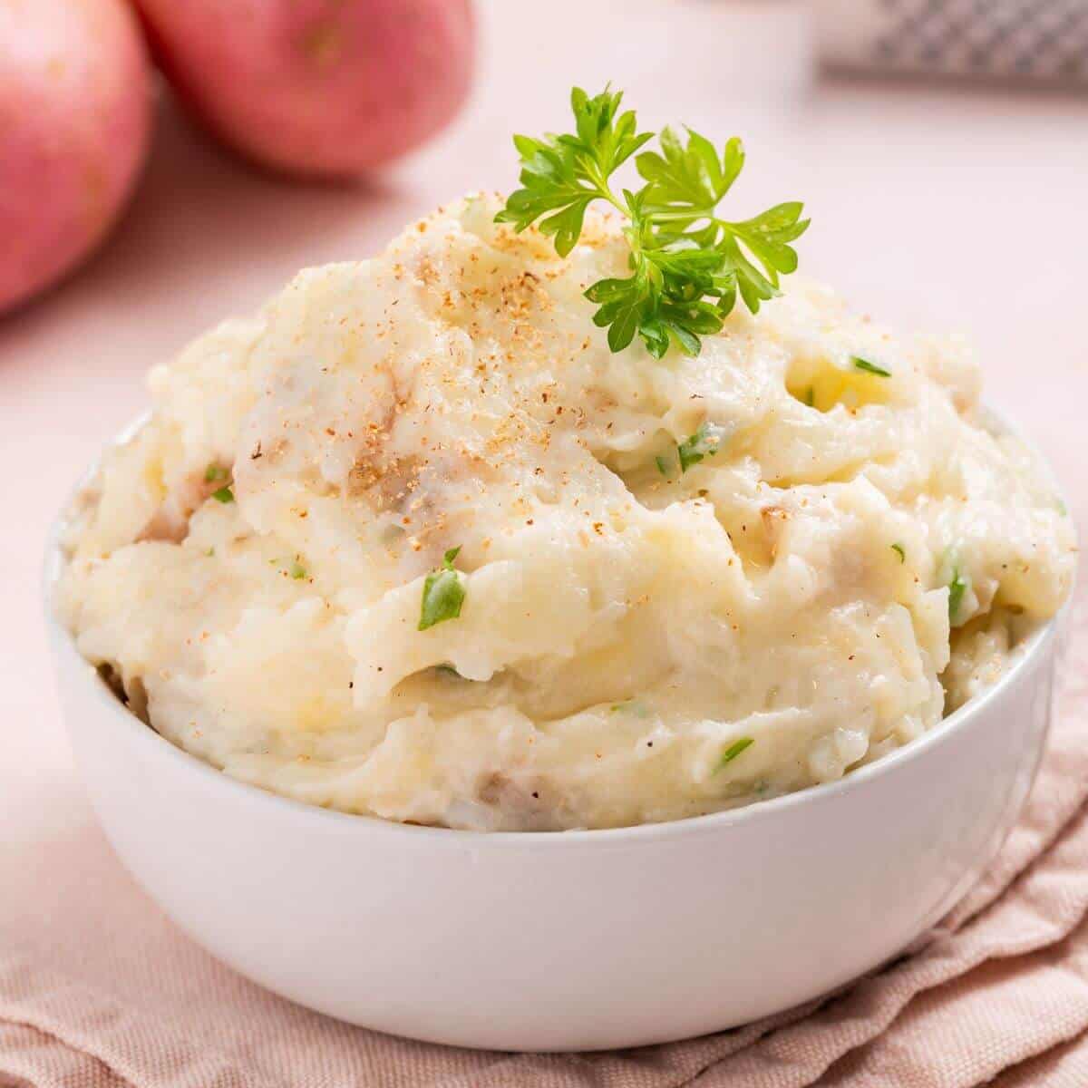 A bowl of mashed potatoes garnished with parsley sits on a table. Red potatoes are visible in the background.