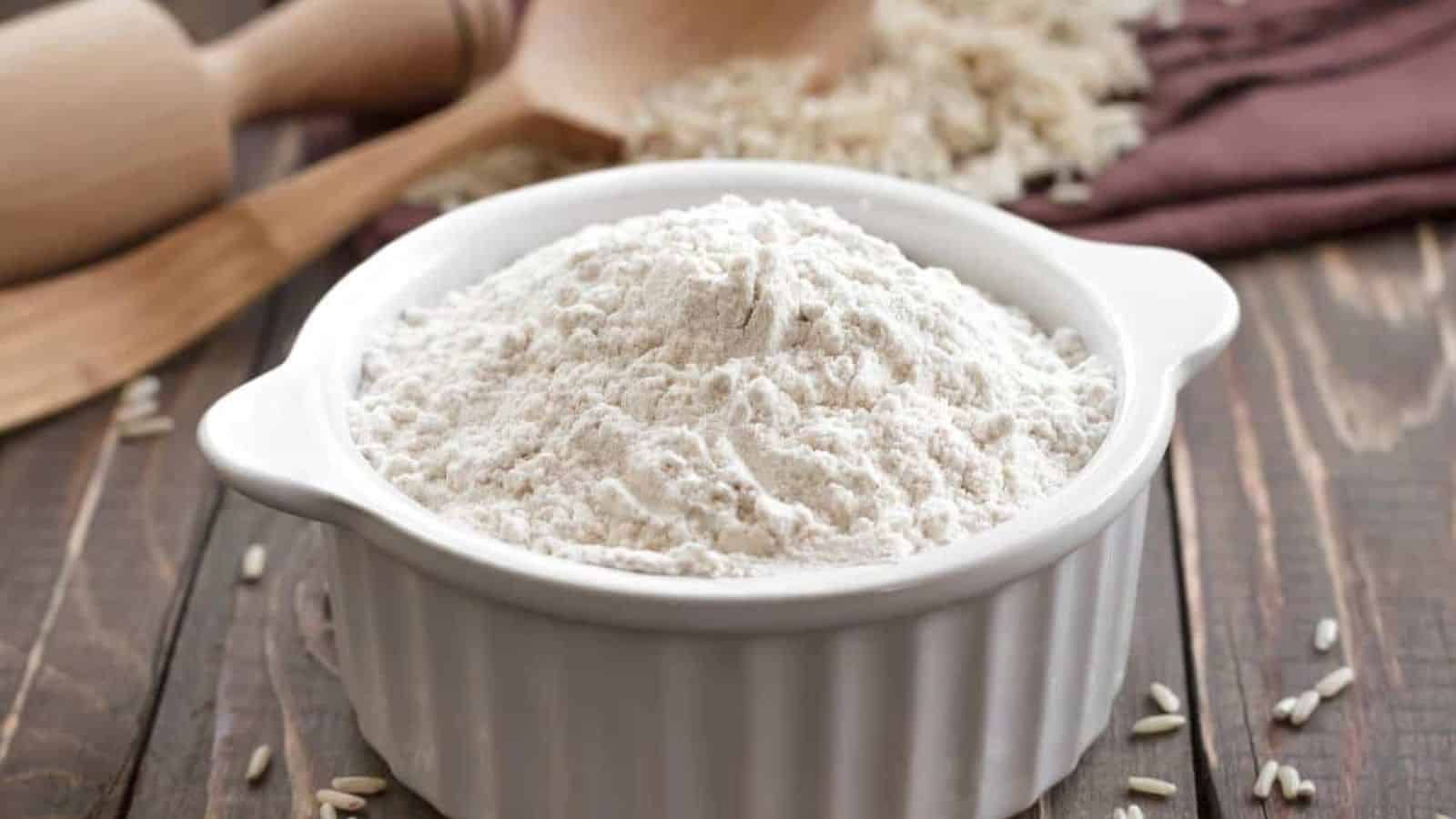 A white ceramic bowl filled with flour, one of those staple foods you should always have in your pantry, sits on a wooden table. It’s accompanied by a wooden spoon and scattered grains.