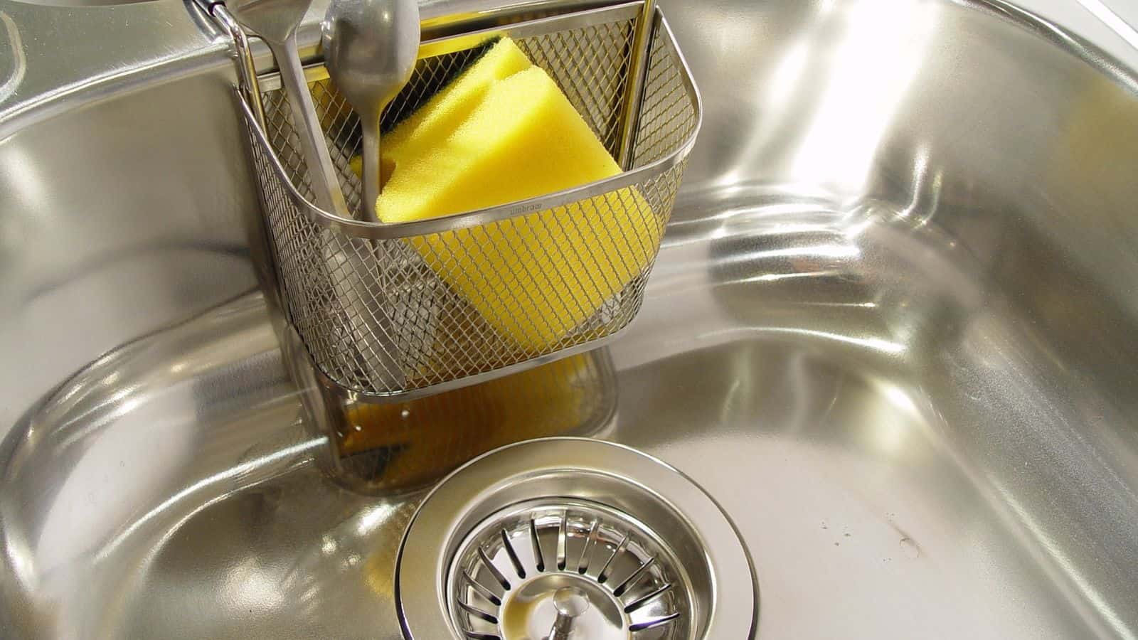 A metal sink with a silver drain has a small metal basket holding a yellow sponge and a spoon.