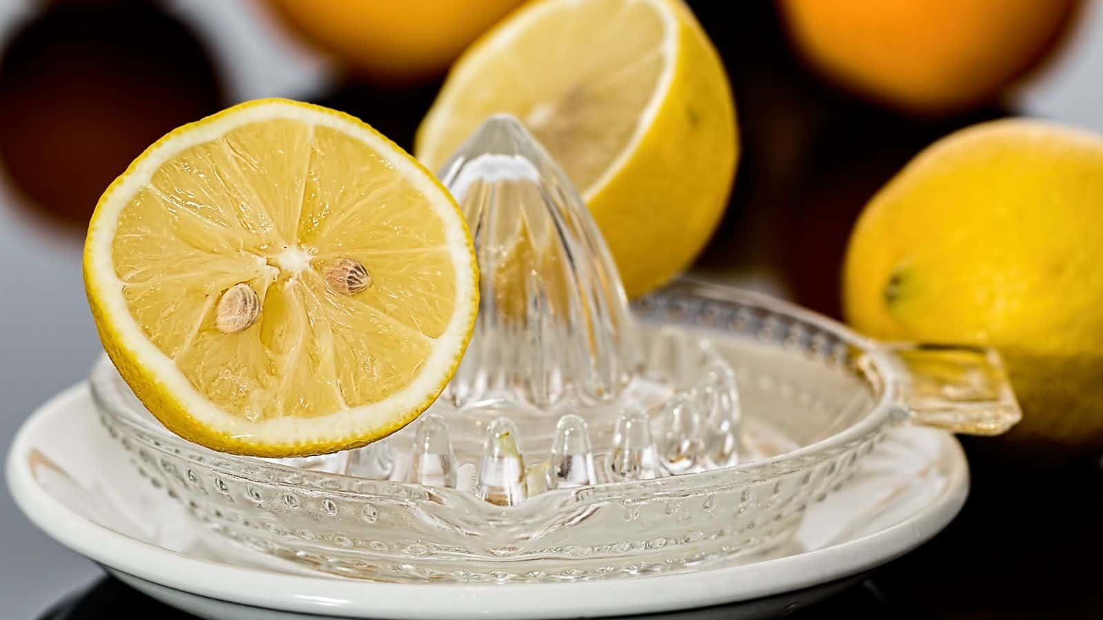 A glass citrus juicer with a sliced lemon and another half lemon next to it. Several whole lemons are in the background, showcasing ingredients that can make your food taste better.