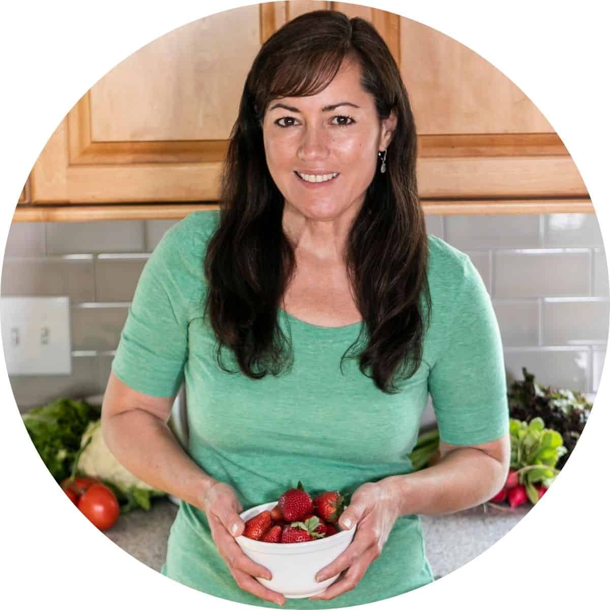 A woman in a green shirt stands in a kitchen holding a bowl of strawberries.