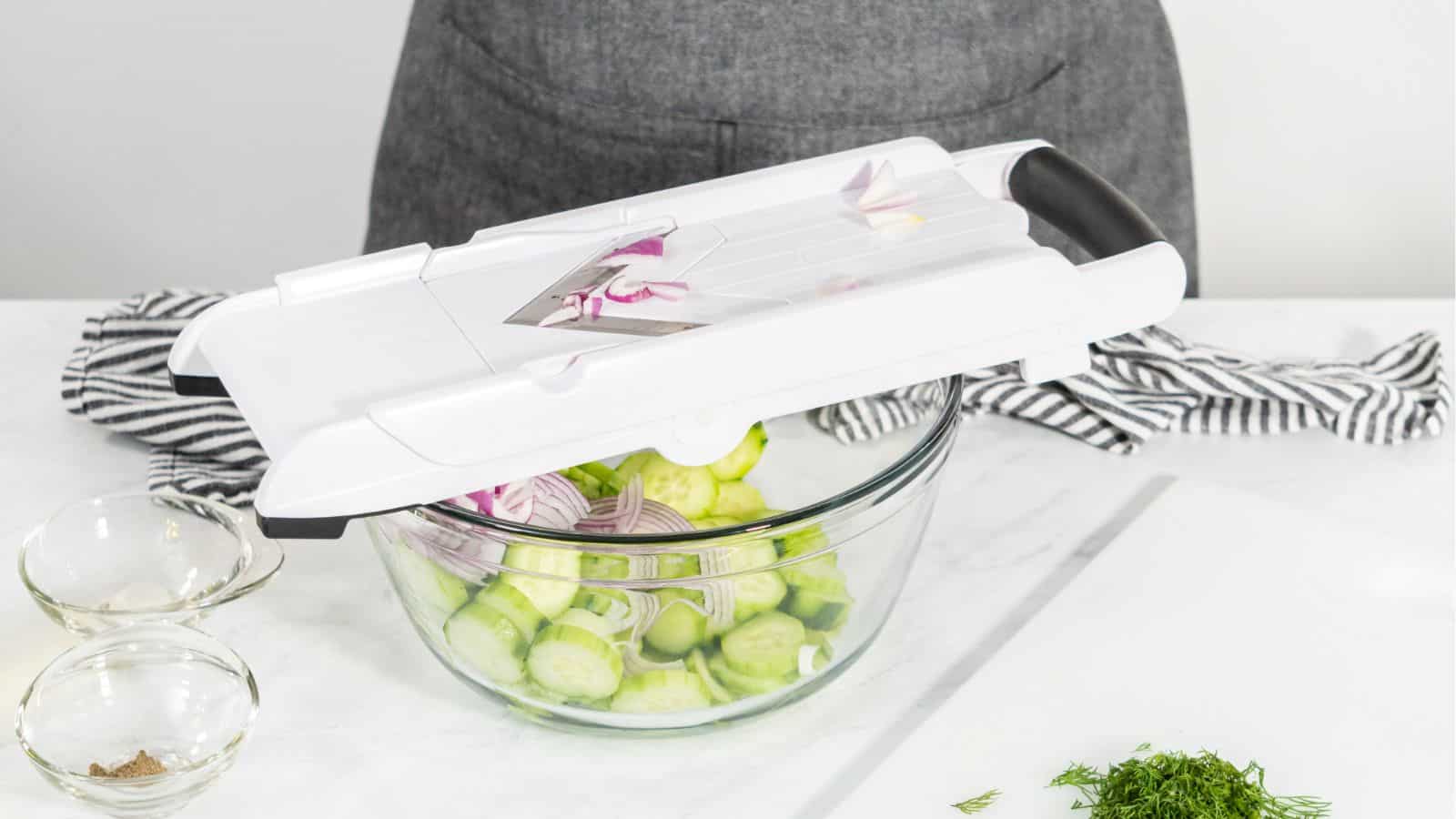 Person slicing cucumbers and red onions into a glass bowl using a mandoline slicer, next to two small bowls and some herbs on a white countertop.