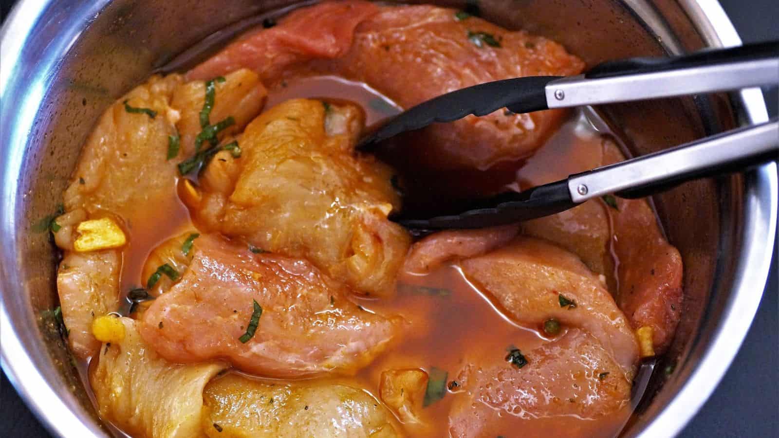 A bowl of raw marinated chicken slices with herbs and garlic, being stirred with metal tongs.