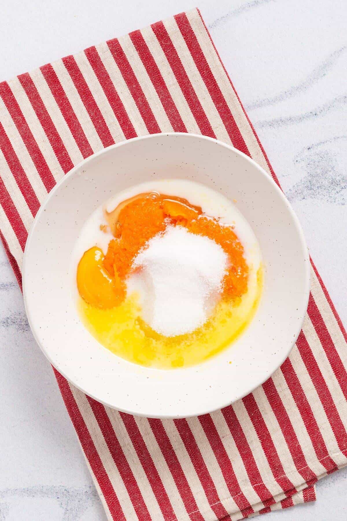 A white bowl containing egg, pumpkin, sugar, and milk is placed on a red and white striped cloth on a white surface.
