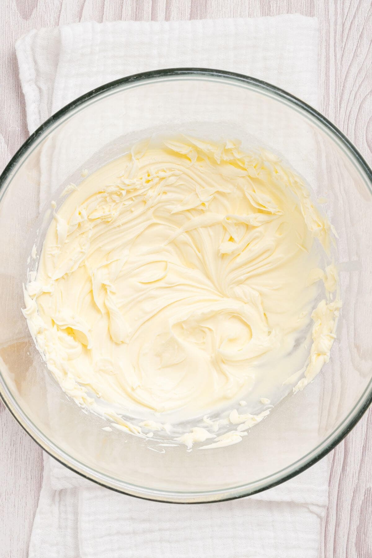A glass bowl containing evenly mixed, creamy, pale yellow butter mixture placed on a white cloth napkin over a light wooden surface.