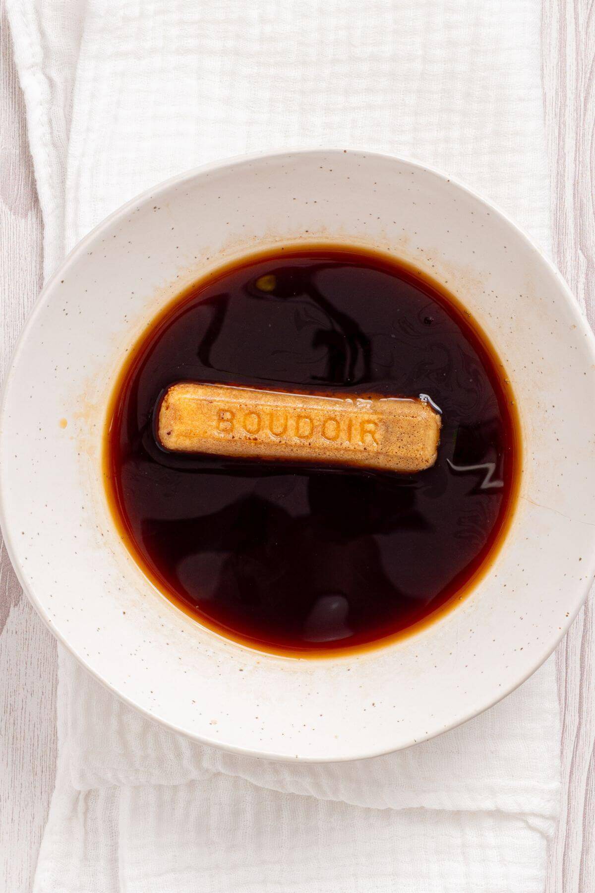 A ladyfinger biscuit labeled "BOUDOIR" is partially submerged in a dark liquid, likely coffee or cocoa, in a speckled white bowl placed on a white textured cloth.
