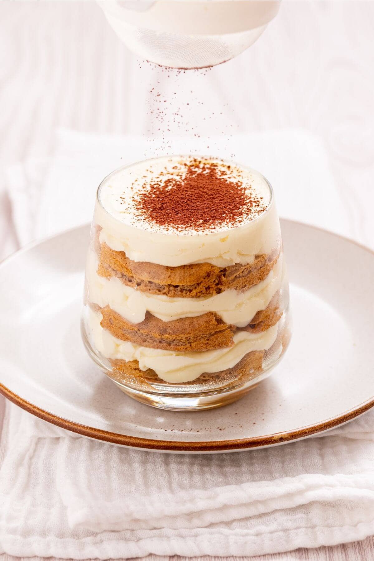 A glass cup filled with layered tiramisu sits on a white plate. Cocoa powder is being sprinkled on top.
